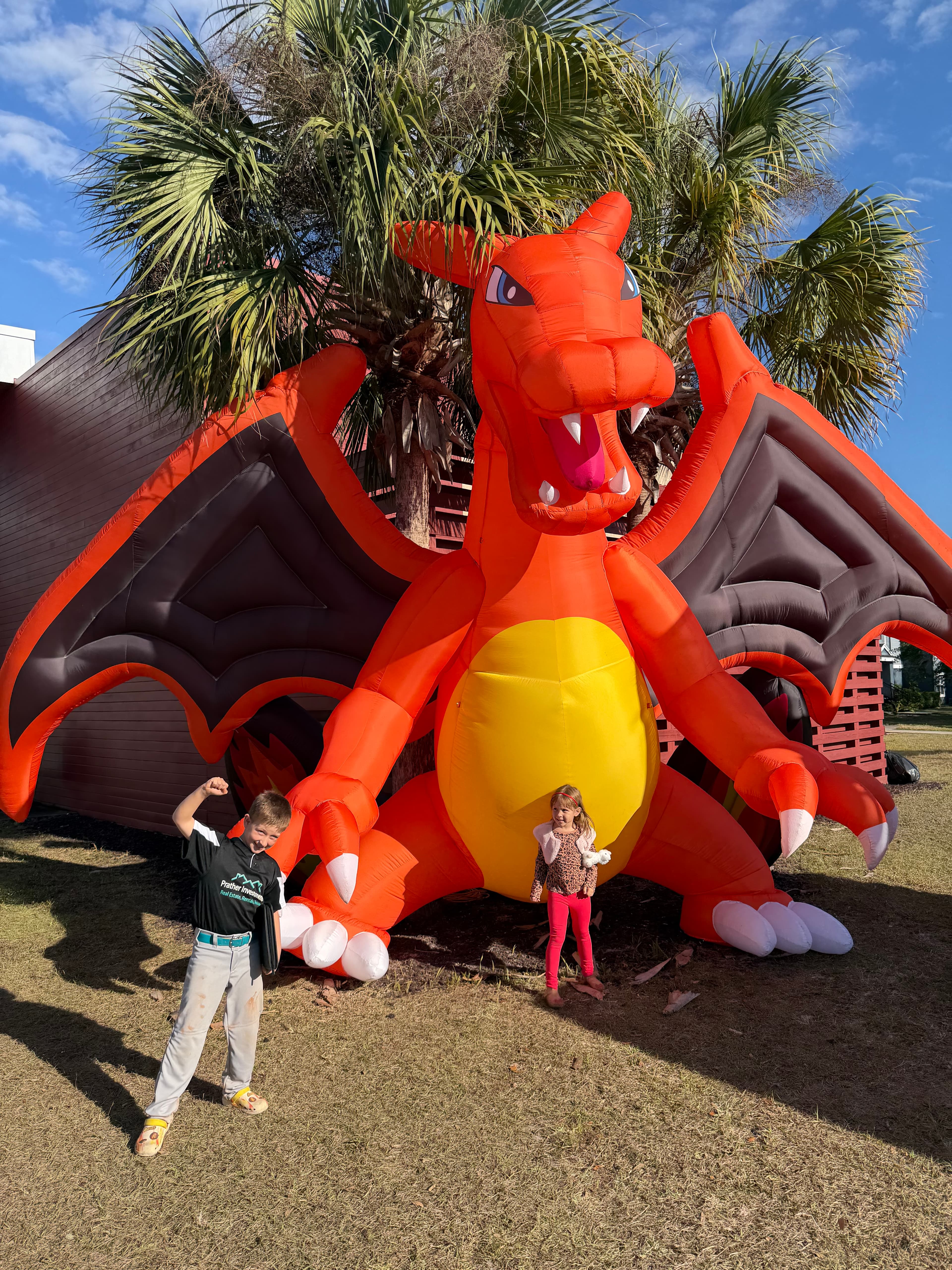 Two children pose with a giant orange inflatable Charizard dragon on a sunny lawn.
