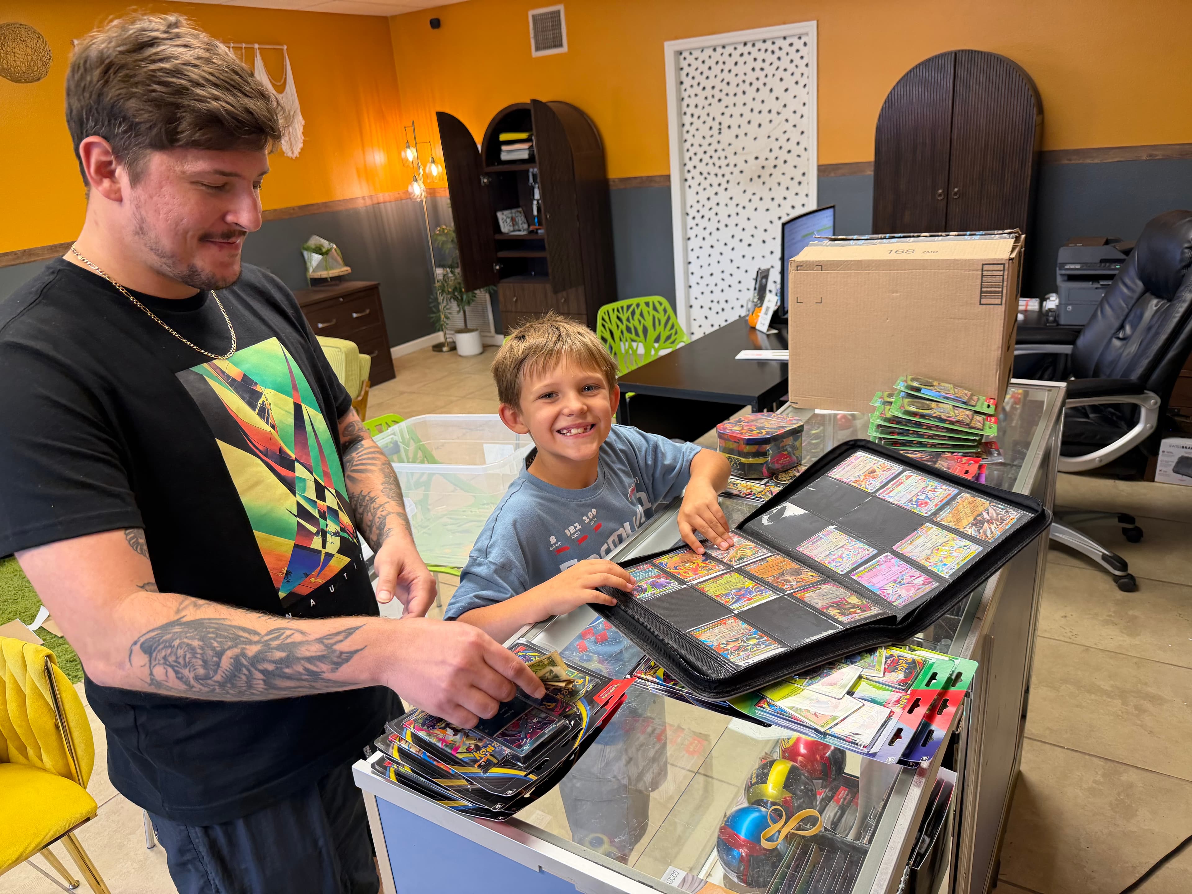 A tattooed man and a smiling boy look at a binder of trading cards.