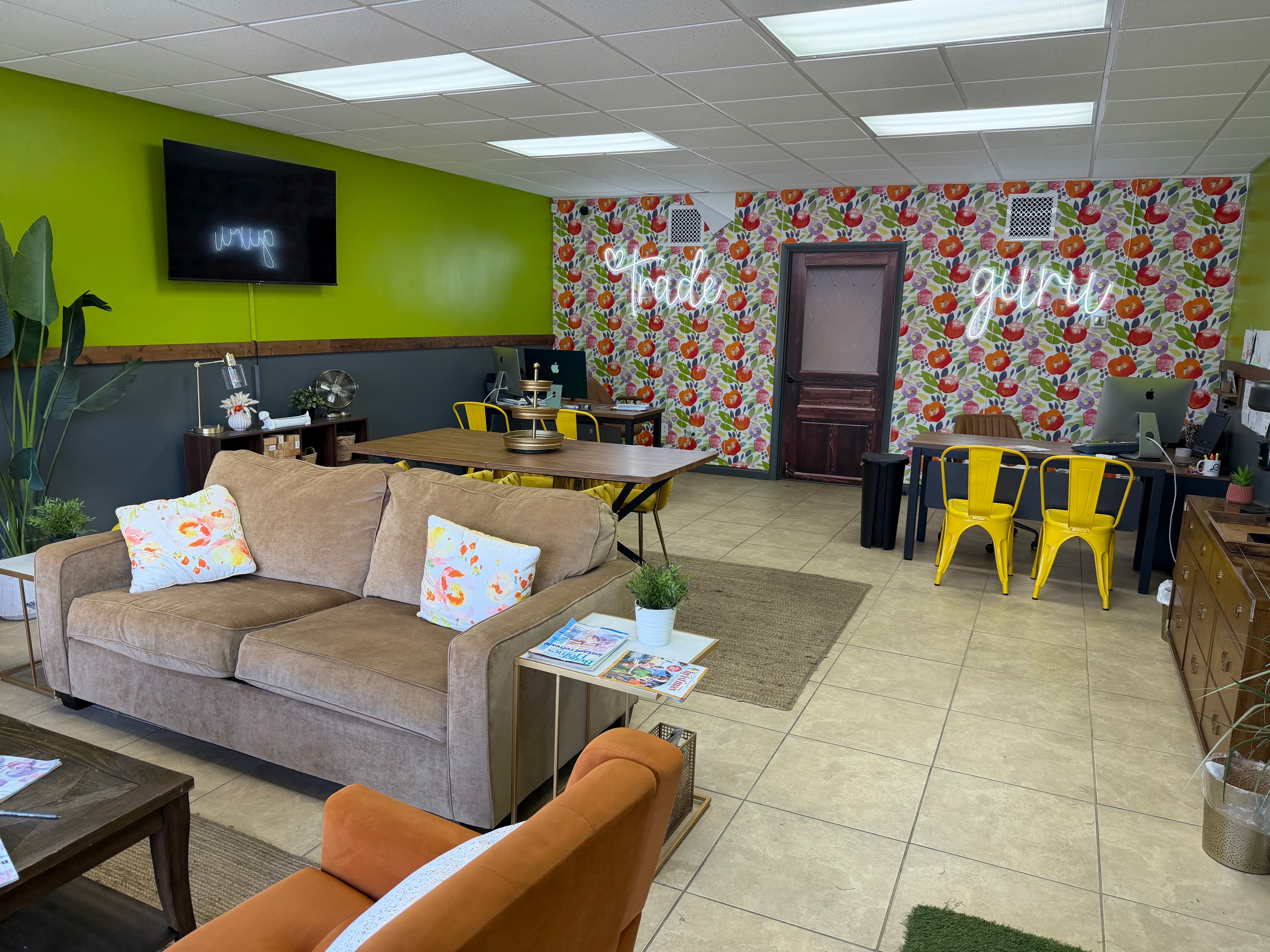 Modern office lounge with a tan sofa, floral wallpaper, neon signs, and yellow chairs.