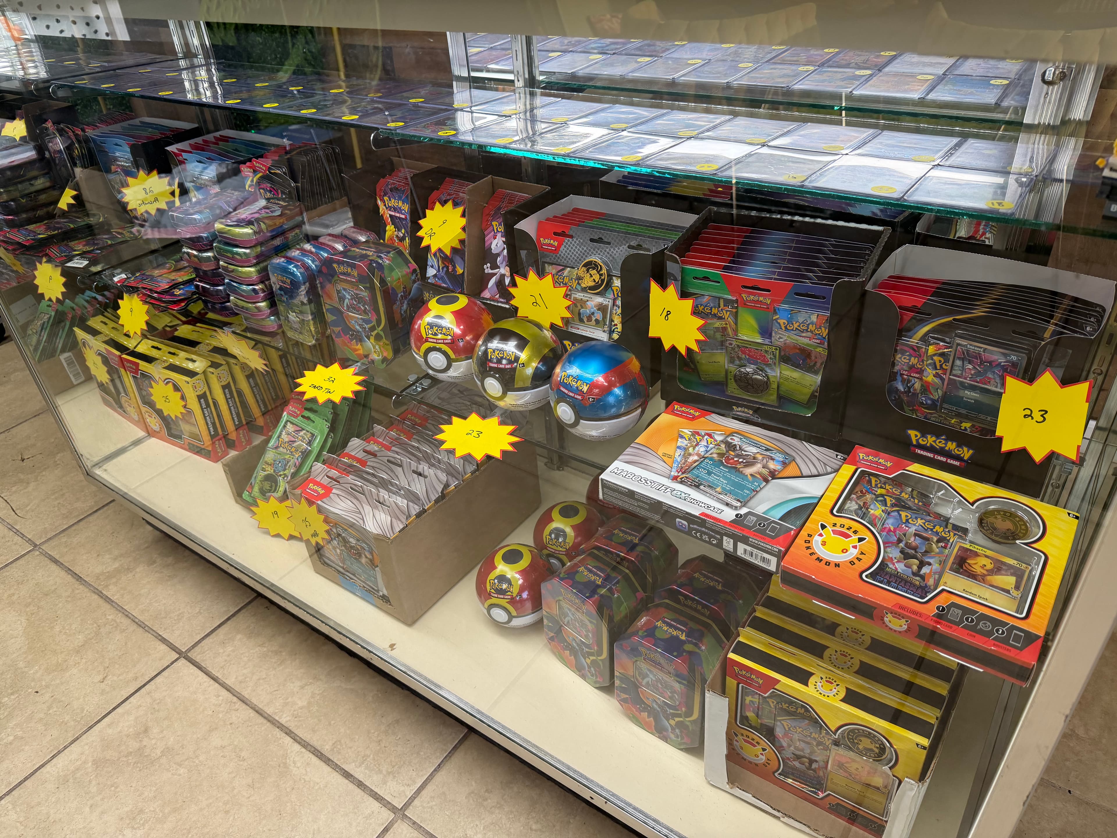 Glass display case filled with Pokémon trading card game tins, booster packs, and Poké Balls.