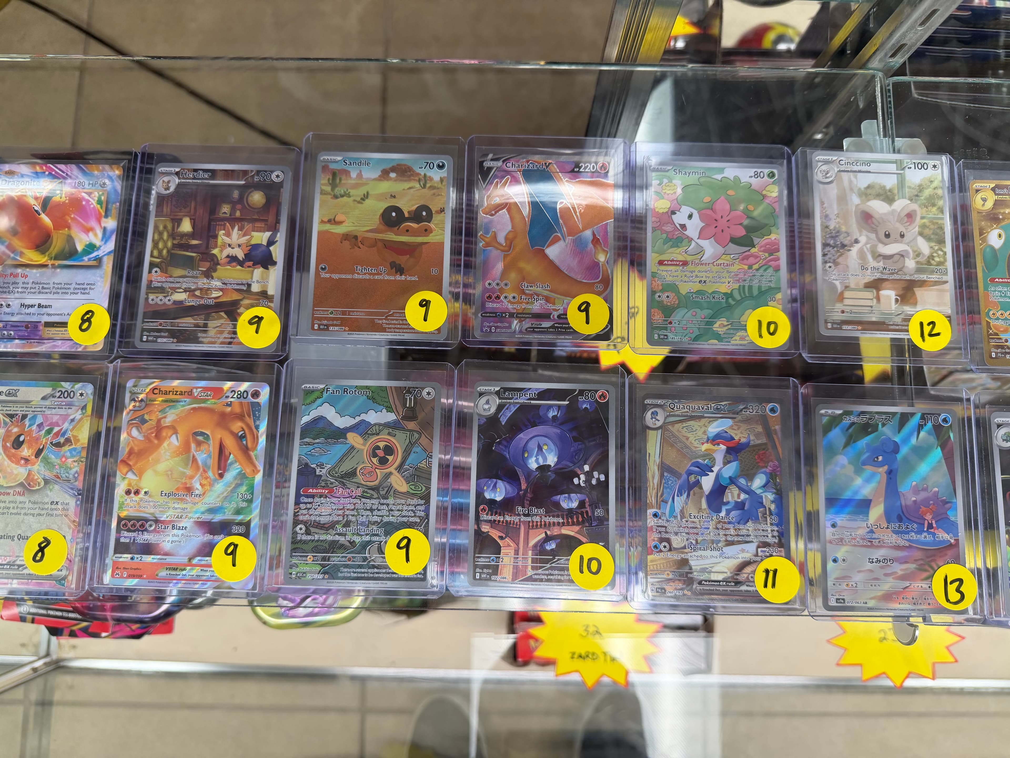 Pokémon trading cards in protective sleeves with yellow numbered stickers displayed in a glass case.